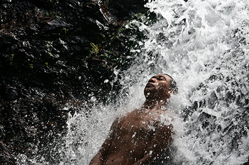 A male swimmer leans back into a Samoan waterfall with a blissful expression on his face