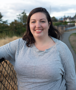 Instructor and founder Naomi Tomky, a Caucasian woman in a grey shirt, smiling as she leans against a fence in the Pacific Northwest. A curving trail and evergreen trees are visible in the background