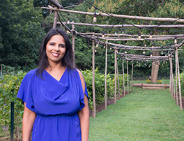 A smiling Indian-American author, wearing an elegant purple dress, against a garden backdrop with a trellis of lights