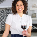 Food writing instructor Kae Lani Palmisano, a smiling biracial woman with dark shoulder-length curls. She's posed in a chic kitchen, holding a glass of red wine and wearing a white, short-sleeved blouse with a pussycat bow.