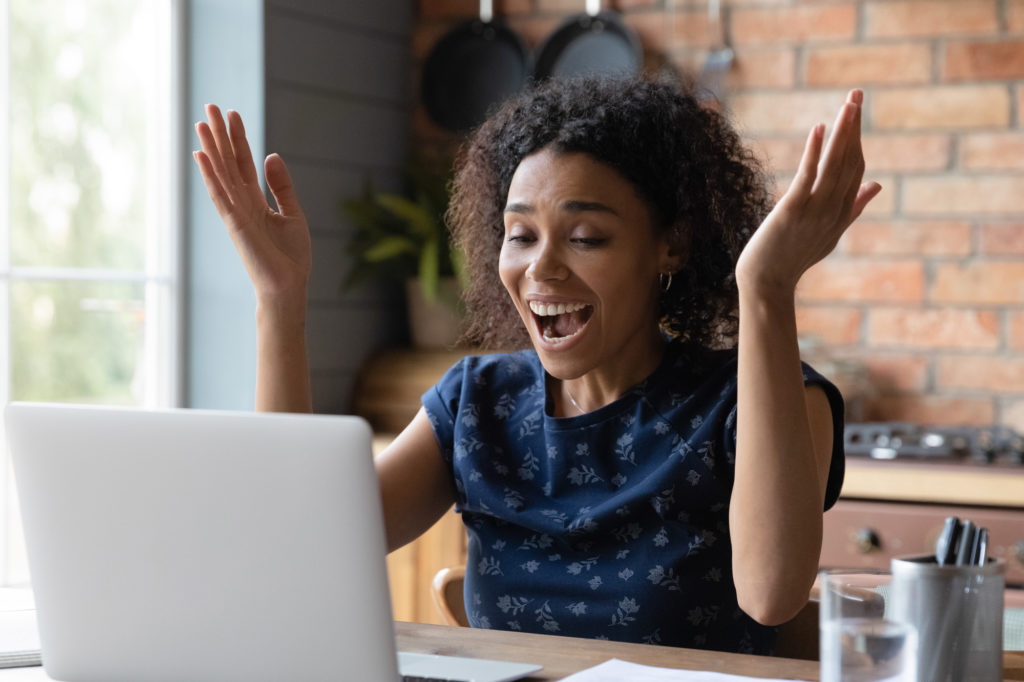 A woman raises her hands joyfully, while gazing at her open laptop. She has brown skin and loose dark curls to her shoulders, and is wearing a navy short-sleeved top. She appears to be working in a kitchen with pans hanging on a brick wall behind her.