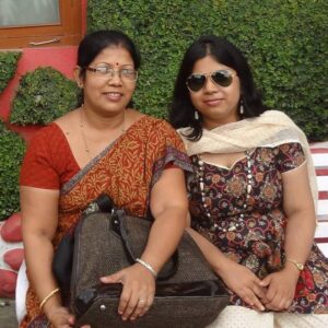 Two Indian women: Tripti Bhuyan and her daughter Arundhati Nath. Mrs Bhuyan is wearing an orange sari, bindi, a low ponytail or bun, and glasses with clear frames. Her shoulder is pressed against her daughter's — and Arundhati has long black hair, black sunglasses, a cream scarf, and sundress. They're sitting in front of what appears to be a hedge